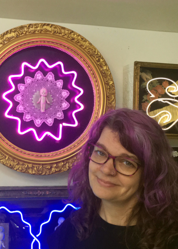 Headshot of Eve Hoyt in front of a white wall with neon art pieces arranged on it. Eve has shoulder-length hair and glasses. Three neon pieces are featured in the background inside of gold frames. The piece on the bottom left shows two neon blue ruffly strands that intersect in the middle. Only a portion of this piece was captured on camera. Above this piece, is a circular gold frame with with a purple neon circle that has points instead of being smooth. Inside this circle is a purple lacey circular piece with points that complement the points on the neon circle. In the center of this circle is a small baby doll. This piece reflects purple into Eve's hair. The piece on the right, next to the purple, is only partially captured by the camera but features strands of white neon on a dark green background with light red flowers and light green leaves.