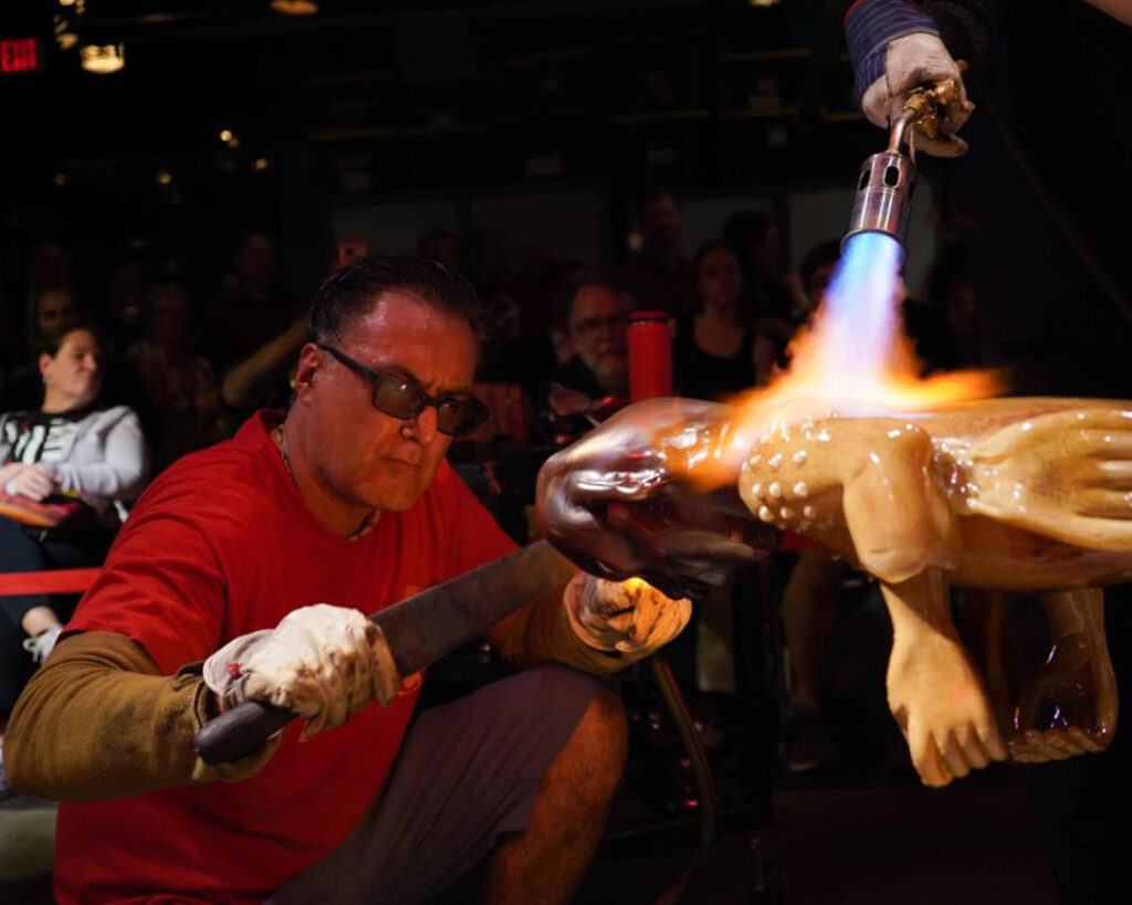 Image of WheatonArts 2024 Creative Glass Fellow Jaime Guerrero using a large metal paddle tool and a torch on a large human-shaped glass piece. in front of a crowd of viewers. Another artist is using a torch on piece as well in the top right corner.