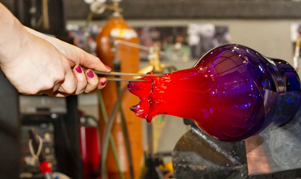 A close up image of vase-shaped hot glass in the WheatonArts Glass Studio. The glass starts dark blue at the bottom, turns purple, and then red at the top. A hand extends from the top left corner of the image and is holding a large pair of metal tweezers. The hand is using the tool to pull and shape the red hot glass forming a ruffle-like pattern around the rim of the vase.