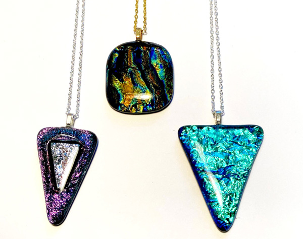 Image of three fused glass pendants resting on a white background. The pendant on the left is a sparkly purple and blue color, a triangular shape with a smaller triangle in the middle, and attached to a silver chain. The pendant in the middle is square shaped with rounded corners, shades of blue and gold in a marbled style, and attached to a gold chain. The pendant on the right is triangular, light blue with dark blue texture, and attached to a silver chain.