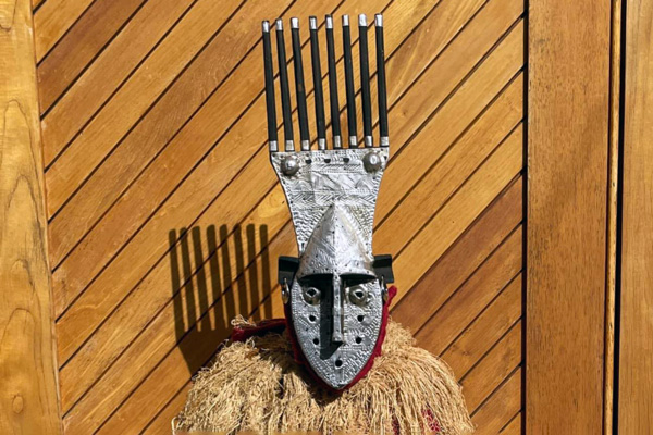 Image of a piece from the WheatonArts Down Jersey Folklife Center. The piece is in front of a diagonal wood background and is comprised of a straw-like upper body, a round diamond-shape silver mask, another rectangular piece of silver that curves in on the sides on top of the mask, and eight tall black tubes standing on top of the rectangular silver piece. Each tube has a thick silver stripe around the top and bottom it. The mask has two eyes, a pronounced nose and forehead, five holes that loop around the nose, and a black rectangular piece on each side of the forehead that connect to a red strip that goes around the bottom of the mask.