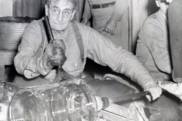 Black and white image of Charles Pepper at Whitall Tatum. Charles is sitting at a bench and is holding a glass blowing rod with his right hand. He is wearing a large glove on his left hand, which is using a large sculpting tool on a large jug-like piece of hot glass at the end of the metal rod. Charles is wearing glasses, a long sleeve shirt, and suspenders. Other people are partially captured in the photo in the background.