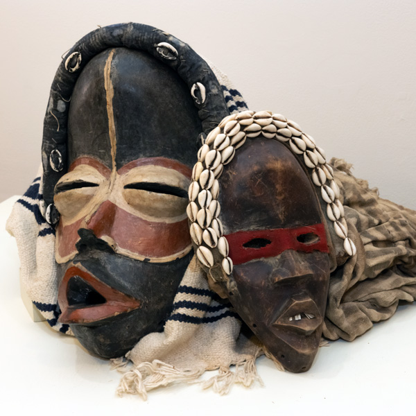 Image of two masks resting on a white to light gray gradient backdrop. The mask on the left is black with red lips and a round red mask around the the eyes and nose. Around the eyes is smaller beige round mask. There is more beige outlining the bottom of the red mask. There is also a matching vertical stripe that starts at the top of the entire black mask and extends through the middle of the forehead to the beige part of the red mask stopping right before the red mask ends around the nose. Around the top of the black mask is a long cylindrical black head piece with six small white cowrie shells along the front side of it. A white cloth with fringe and three dark blue stripes towards the top and middle of it rests around the entire mask. The mask on the right side of the image is smaller than the one on the left. This mask is black with light brown highlights and speckles throughout the base. This mask has white teeth with a gap in the center and a thin, rectangular red mask around the eyes. This mask also has a cylindrical head piece draped around the top of it. This piece is the same color as the rest of the base of the mask, but is covered in three rows of small white cowrie shells lined up next to one another alongside the top and front side of the head piece. Behind this mask is another cloth. This cloth has a pattern of gray and beige stripes.