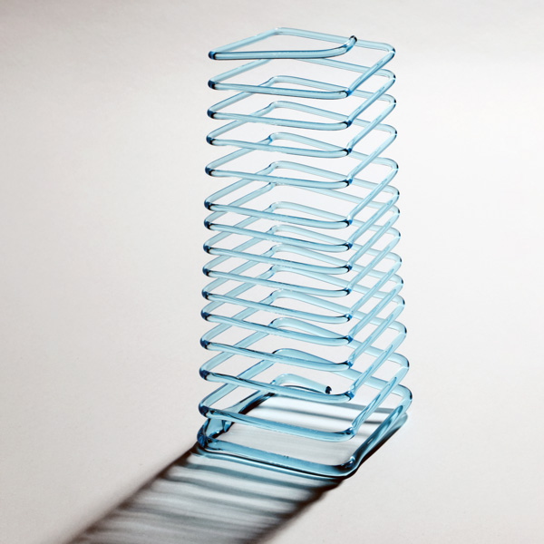 Image of a glass Jacob's Ladder found in the WheatonArts Museum of American Glass. The tall piece is made up of a long continuous thin glass rod that spirals in square shapes from top to bottom. The piece is resting on a white background and it's shadow extends from the piece to the bottom left corner of the image.