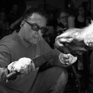 Black and white image of WheatonArts 2024 Creative Glass Fellow Jaime Guerrero using a large metal paddle tool and a torch on a large human-shaped glass piece. in front of a crowd of viewers. Another artist is also using a torch on piece in the top right corner.