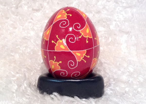 Image of a painted egg on top of a circular black stand resting on and in front of a soft white backdrop. The egg has been painted red with small white vertical lines dividing the egg into even sections. There is also another thin white line around the middle of the egg. In the center of these lines extend two orange three-petal flowers with yellow centers and two yellow stamens. There is one flower on one side of each line as they mirror one another. Each flower is outlined in white and has a swirly white line extending from the middle where the flowers meet.