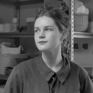 Grayscale headshot of WheatonArts 2024 Creative Glass Fellow Lily Wilkins. Lily is wearing a long-sleeve shirt with a collar, buttons, and a pocket on the right side. Lily is looking off to the side in the image and her hair is tied back.