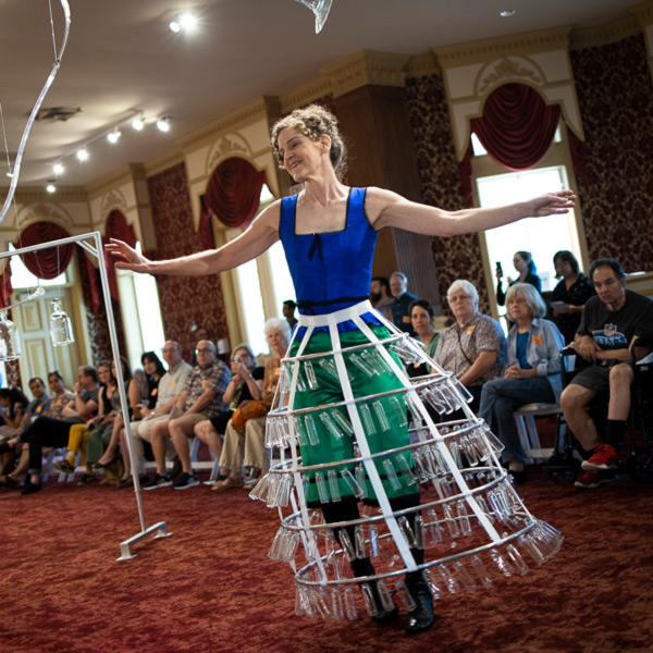 Image of a performance in the lobby of the WheatonArts Museum of American Glass. The dancer is wearing a blue corset top with a small black bow on the chest and another tied around the waist. The dancer also has teal pants, long black boots, and a white pannier of a five-tier hoop skirt. This piece has small clear glass tubes dangling around each of the hoops. The dancer has both arms out to help move in a way that allows the skirt to make noise. A crowd of people sit in the background watching the performance. Also in the background is a large white rack with more clear glass hanging from it and portions of other clear glass pieces are at the top of the image. Each glass piece was used in part of the performance.