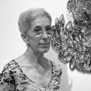 Black and white headshot of WheatonArts 2024 Creative Glass Fellow Nancy Cohen. Nancy has short hair, glasses, and a shirt with dark speckles. Nancy is standing in front of a white wall with a portion of a piece of artwork pictured on the right side of the image.