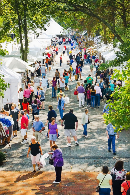 Image of a crowd outside at WheatonArts. The crowd is walking through a paved lane with white tents on each side. There are shoppers gathered inside and in front of the tents.