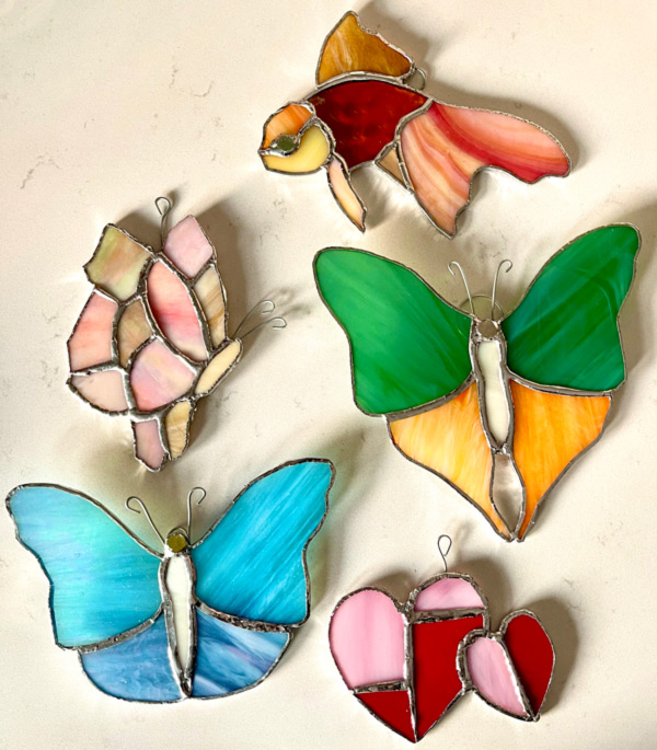 Image of five stained glass pieces resting on a white a background. There are two pieces at the bottom of the image, two in the middle, and one at the top. The bottom left piece is a stained glass butterfly with blue wings, a white body, and a gold head. The piece on the bottom right is comprised of two pink and red hearts. The heart on the left is larger than the one on the right. The piece above the hearts is another butterfly with green top wings and yellow bottom wings. The body of this butterfly is white and it has a silver head. The piece next to this one is the side profile of a butterfly. The wings are a mix of pink and a light beige color. The body is also this beige color. At the top of the image is a piece that is in the shape of a fish. The fish has a yellow head and fins, a red body, and a long tail that is white with red and orange striped throughout.