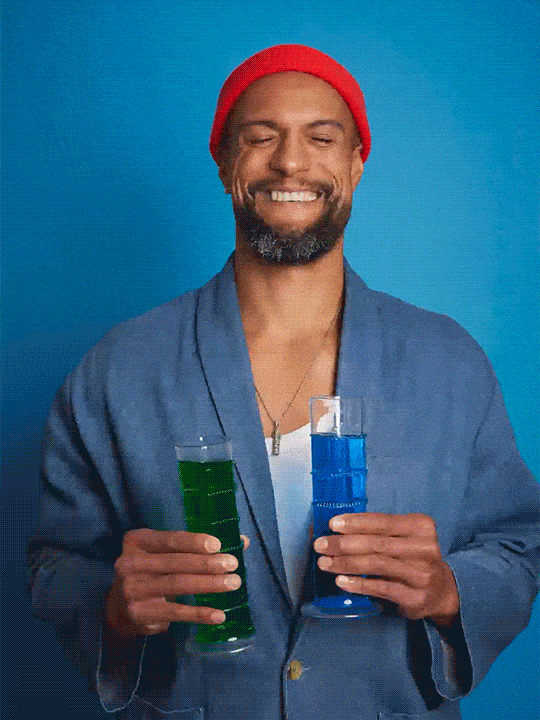 Gif of a person with a large clear graduated cylinder in each hand. The cylinder on the left is filled with green liquid and the cylinder on the right is filled with blue liquid. The gif shows this person holding each cylinder up towards their face as they consider the contents. The person then takes a sip of the liquid. This repeats with the cylinder on the right. The person smiles at the camera before the gif loops back to the beginning. The person has a short beard and is standing in front of a blue wall and is wearing a blue blazers over a white shirt, a necklace, and a red beanie.
