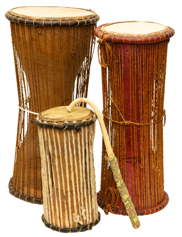Image of three pieces from the Ceremonies in Circles exhibit on a white background. There are two tall drums that are wide at the ends and thin in the middles. The drum on the left has a light brown base with light brown stings extended vertically from the top of the drum to the bottom. A white strand is tied around the middle. The drum on the right is slightly shorter than the one on the left with a light brown strand tied around the middle and a reddish base color. In front of them is a smaller drum about half the size of the taller ones. This drum has a light beige base color and white vertical strands. Resting on top of the drum is the curved portion of a long, thin light beige piece with green patterned fabric wrapped around the end.