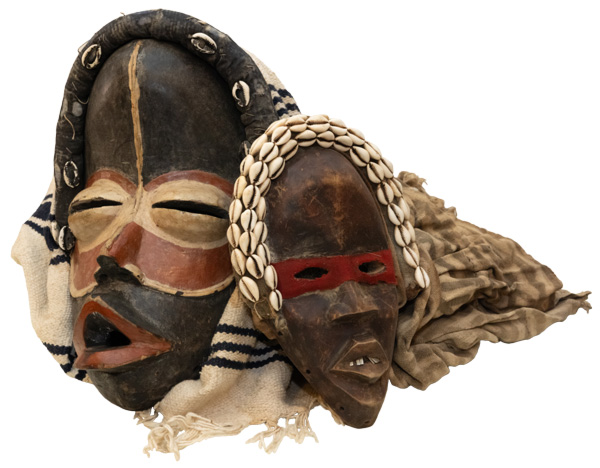 Image of two masks resting on a white backdrop. The mask on the left is black with red lips and a round red mask around the the eyes and nose. Around the eyes is smaller beige round mask. There is more beige outlining the bottom of the red mask. There is also a matching vertical stripe that starts at the top of the entire black mask and extends through the middle of the forehead to the beige part of the red mask stopping right before the red mask ends around the nose. Around the top of the black mask is a long cylindrical black head piece with six small white cowrie shells along the front side of it. A white cloth with fringe and three dark blue stripes towards the top and middle of it rests around the entire mask. The mask on the right side of the image is smaller than the one on the left. This mask is black with light brown highlights and speckles throughout the base. This mask has white teeth with a gap in the center and a thin, rectangular red mask around the eyes. This mask also has a cylindrical head piece draped around the top of it. This piece is the same color as the rest of the base of the mask, but is covered in three rows of small white cowrie shells lined up next to one another alongside the top and front side of the head piece. Behind this mask is another cloth. This cloth has a pattern of gray and beige stripes.