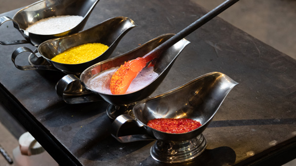 Image of four metal containers of glass frit on a small metal table. From left to right, the frit colors are white, yellow, white, and red. A metal rod with a small mass of orange hot glass at the end of it is being dipped into the white frit that is second from the right of the line of containers. Pieces of frit are sticking to the hot glass.