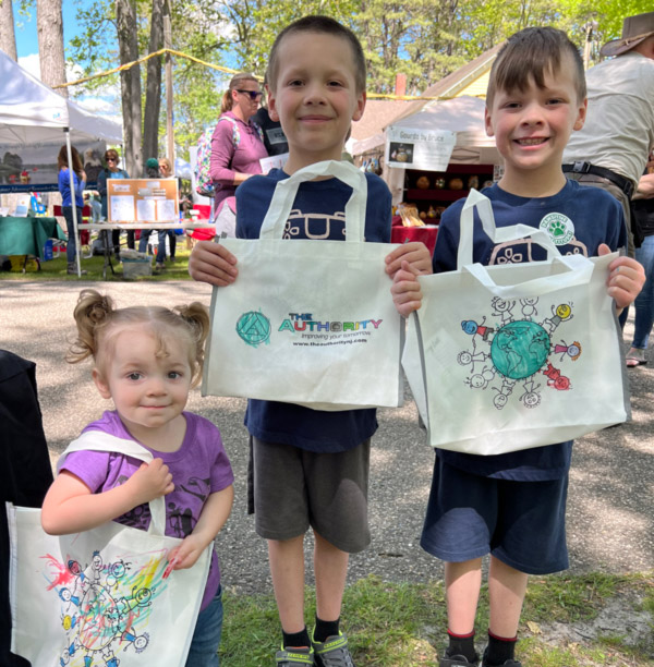 Image of three children outside at WheatonArts during Eco Fair posing with their tote bags for the camera. The child on the right has a white tote bag with cartoon drawings of children standing around a drawing of the Earth. The drawings have been colored with multicolored scribbles. The child in the middle's white tote bag has a smaller cartoon Earth next to colorful block letters that read "The Authority". The child on the right's tote bag is similar to the child on the left's with parts of the drawings colored in with colorful colors. There are Eco Fair tents and tables in the background.