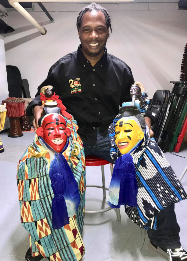 Image of Ira L. Bond sitting on a red stool holding a piece of artwork in each hand while smiling at the camera. The artwork on the left side is a red mask with a orange, beige, and blue patterned cloth wrapped around the bottom of it. At the top of the mask is a small sculpture of a person. The mask has a white smile with black eyes, eyebrows, a vertical strip of black on the forehead and two horizontal stripes on the cheeks. The artwork on the right side is similar to the one on the left, but is a yellow mask with a blue, black, and white patterned cloth wrapped around it.