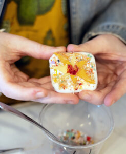 Image of a square-shaped fused glass make your own piece held with two hands towards the camera. Under the piece is a clear plastic cup with colorful glass frit inside of it and a silver utensil. The base of the piece is white. It has red frit gathered in a small circle in the middle of the piece, seven yellow triangular pieces gathered around the red, and small multicolored frit pieces gathered around the design in the background on top of the white base.