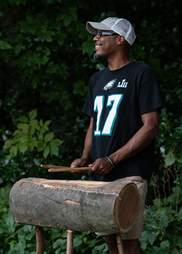 Image of Richard Robinson outside in front of a green leafy background from the side. Richard is standing behind a log held up by three tube-shaped stands and is holding a drum stick in each hand above the log.