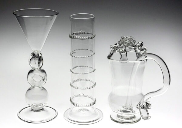 Image of three clear glass pieces from the Centuries of Tomfoolery exhibit in front of a dark gray-to-white gradient background. The background starts white at the bottom and gets darker towards the top. The piece on the left is a tall clear glass that has a circular disc at the bottom, three sphere like shapes as the stem, and a funnel-shaped top. The sphere in the center has a small circular cut-out in the middle of it. The glass piece in the center is clear and shaped like a tall graduated cylinder with five thin textured circles evenly spaced throughout the cylinder. The clear piece on the right is shaped like a small pitcher with a long handle that starts inside the glass and extends until the bottom of the piece. Three small clear spheres rest at the bottom of the curvy handle. At the top of the piece are four pieces of glass evenly spaced around the rim. These pieces are holding a round, curvy clear glass piece at over the opening of the pitcher.