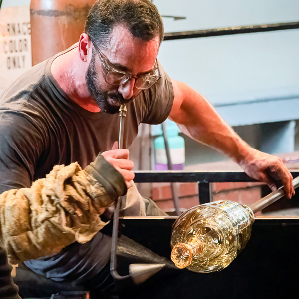 Image of WheatonArts Glass Artist Alexander Rosenberg working at a glass blowing bench. Alex is using one hand on a glass blowing rod that has a large yellow vessel-shaped hot glass piece with a smaller tube-like opening at the top of it towards the end of the rod. Alex is blowing into a metal tube that curves at a right angle towards the hot glass. There is a small cone towards the end of the tube. Alex is wearing glasses, a short sleeve black shirt, and a large protective sleeve on the left arm.