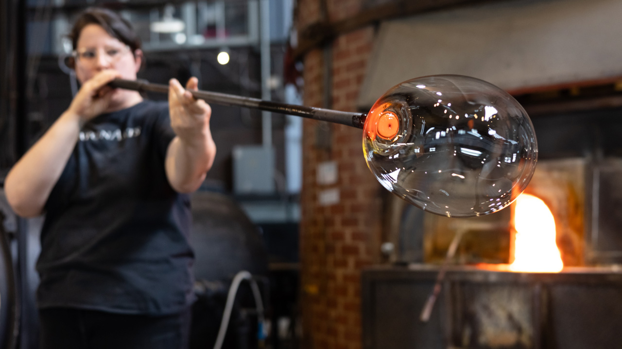 Image of a member of the WheatonArts Glass Studio team blowing into a long metal rod with a large bubble of clear hot glass at the end of it inside of the WheatonArts Glass Studio. The end of the rod before the clear hot glass is bright orange. The team member is wearing clear goggles and a black t-shirt with white text that reads "WheatonArts".
