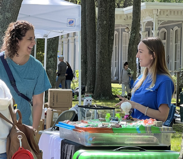 Image taken at Eco Fair outside at WheatonArts in front of the Museum of American Glass. The image shows a visitor smiling and interacting with someone working a booth at the event. The booth is comprised of a table with containers sitting on top of it. The person working the booth is pointing towards one of the containers.