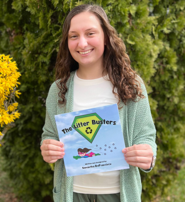 Image of "The Litter Busters" author Samantha DeFrancisco outside in front of a green nature background. Samantha is holding her book and smiling at the camera. The children's book is square shaped with a blue sky cover. In the top center is a diamond shaped logo. The outside of the diamond is a dark green and the center is a light green. Large black text extends across the top of the book through the center of the diamond. This text reads "The Litter Busters" and has a small recycling logo beneath it towards the tip of the diamond. Underneath is a cartoon superhero flying through the sky. The hero has long brown hair, green gloves, a blue long sleeve shirt, a magenta domino eye mask, a magenta cape, a magenta skirt, and green boots. Tiny recycling symbols trail after the hero. More black text is underneath the hero including the authors name. Samantha has long brown hair, a white shirt, and a light green cardigan.