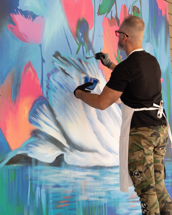 Image of a painter standing in front of a large mural. One hand is using a paint brush to paint a blue streak onto the canvas, while the other hand is holding a small tub of blue paint. The mural is of a large white swan on top of blue water. The background is blue with large pink flowers, green leaves, and dark blue stems. The painter has glasses, a black t-shirt, a white apron, and camouflage pants.