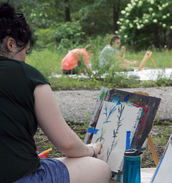 Image of a person sitting and painting outside in nature. The person is sitting on a white cloth in front of a small wooden easel with paint over the front. There is a small rectangular piece of paper taped on each side with blue tape onto the easel. The person is painting leafy images onto the paper. Two more people are in the background sitting on a white cloth and working on their own nature paintings.