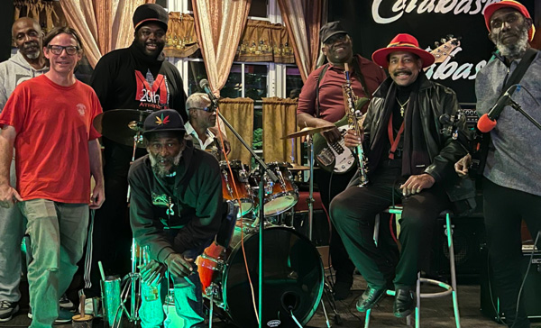 Image of seven members of the Tabernacle Reggae Band posing for the camera on a stage surrounded by instruments and microphones. There are five members on the left side of the image and three on the right. The three members on the right side of the image are holding instruments. Two are holding guitars and the member in the middle is holding a woodwind instrument. There is a large drum set in between the five members and the three.