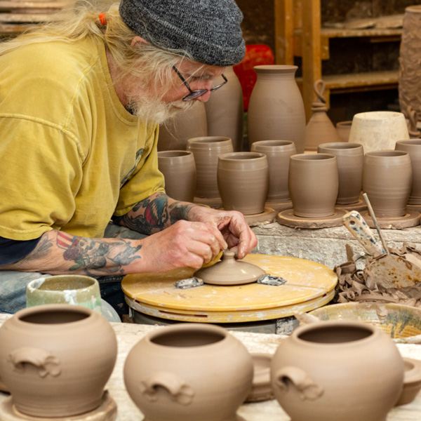 Image of WheatonArts Ceramic Artist Terry Plasket leaning over a tan potters wheel. He is using both hands to sculpt a beige circular lid with a small knob sticking up from the center of it. There are small rectangular silver pieces holding the lid in place. There are three beige clay pots resting at the front of the images, eight beige clay vessels resting on top of beige discs in the background of the photo. Three taller clay vases and the top of a luminary are resting behind the vessels with a few more clay pieces behind them. Terry has a long pony tail and a short beard. He is wearing glasses, a yellow short sleeve shirt, and black short-sleeve shirt under the yellow.