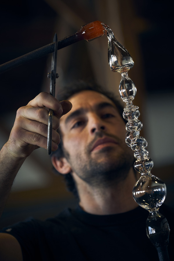 Image of Marc Barreda using a pair of metal tweezers to hold a metal rod steady. The metal rod has a mass of orange hot glass at the end of it and is pulling clear glass at the top of a long, thin clear glass piece that is attached to the end of a different metal rod at the bottom of the image. The piece has been pinched into many different round sections.