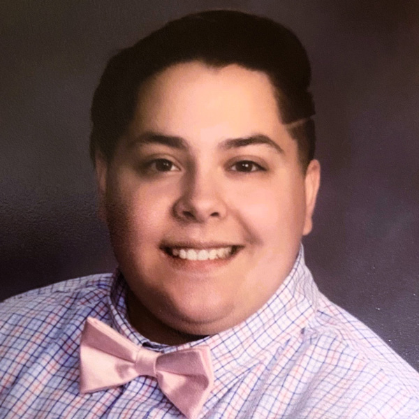 Close up headshot of Marissa Salamoni in front of a dark gray and black backdrop. Marissa has short dark hair and is wearing a light plaid shirt with a light pink bow tie.