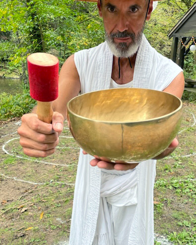 Image of Ken Leap holding a light brown singing bowl in one hand and a wooden mallet with a red top in the other hand. Ken is wearing a long white sleeveless cardigan over a white shirt and white pants. Ken is standing in front of a woodsy area with a white circular labyrinth on the ground in the background. There is also a brown gazebo with yellow string lights on the right side of the image.