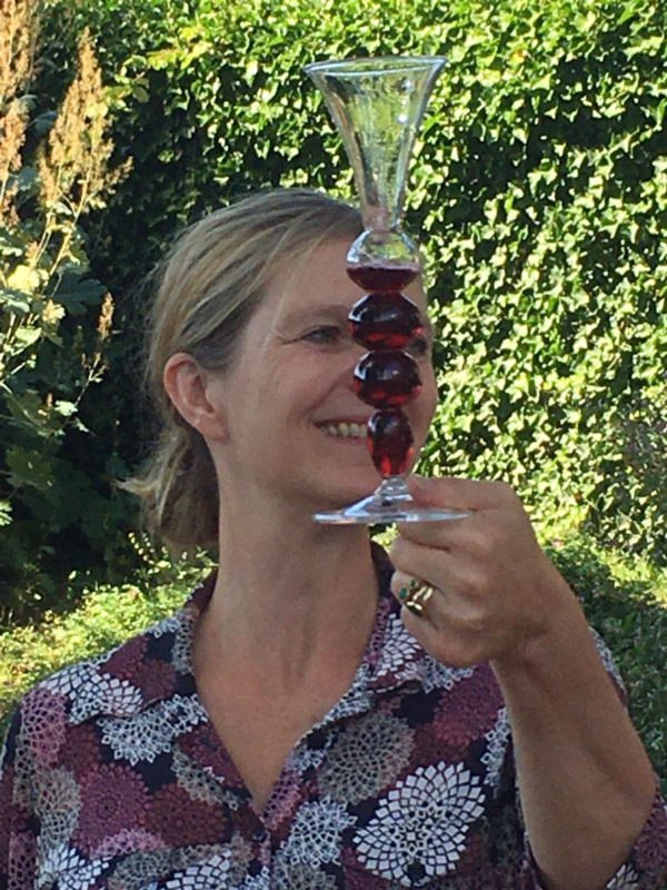 Image of Kitty Laméris outside holding a clear glass by the bottom disc with the right hand. The glass has a bottom disc, four round sections as the stem, and a funnel top. The glass is filled half way up the fourth round section with a red liquid. Kitty has blonde hair tied in a low bun and is wearing a dark red floral button shirt with white, pink, red, and beige mandala-like flowers.