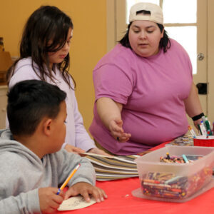 Image of two children at a craft table. One child is sitting at the table and the other is standing. The sitting child is holding a pencil in one hand and is holding a paper steady of the table with the other. The standing child is holding a piece of paper with one hand. An instructor is gesturing towards the paper in the standing child's hand while both children are turned to listen to the instructor's comments. Also on the craft table is a large clear container of crayons and a red cup filled with markers.