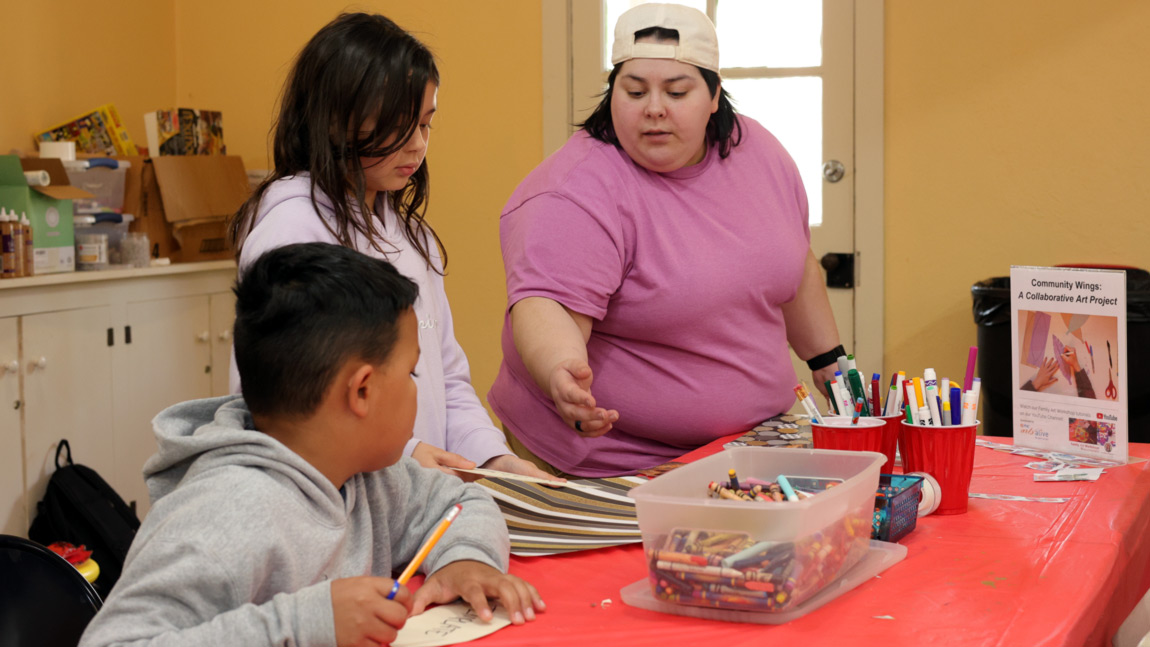 Image of two children at a craft table. One child is sitting at the table and the other is standing. The sitting child is holding a pencil in one hand and is holding a paper steady of the table with the other. The standing child is holding a piece of paper with one hand. An instructor is gesturing towards the paper in the standing child's hand while both children are turned to listen to the instructor's comments. Also on the craft table is a large clear container of crayons, a blue container, three red cups filled with markers, and a sign that reads "Community Wings A Collaborative Art Project".