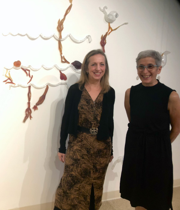 Image of Nancy Cohen and Anna Boothe standing next to one another in front of a white wall with piece of glass artwork arranged on it to create a larger piece. The artwork consists of white curvy tubes of glass and more tubes of red glass extending off of the white. There is also larger red, blue, and white pieces resting on different sections of the larger piece. Anna Boothe has shoulder length light brown hair and is wearing a long brown dress with a black leaf pattern, a black belt with a gold buckle in the center, and a long sleeve black shrug. Nancy Cohen has short silver hair, blue glasses, and is wearing a sleeveless black dress.