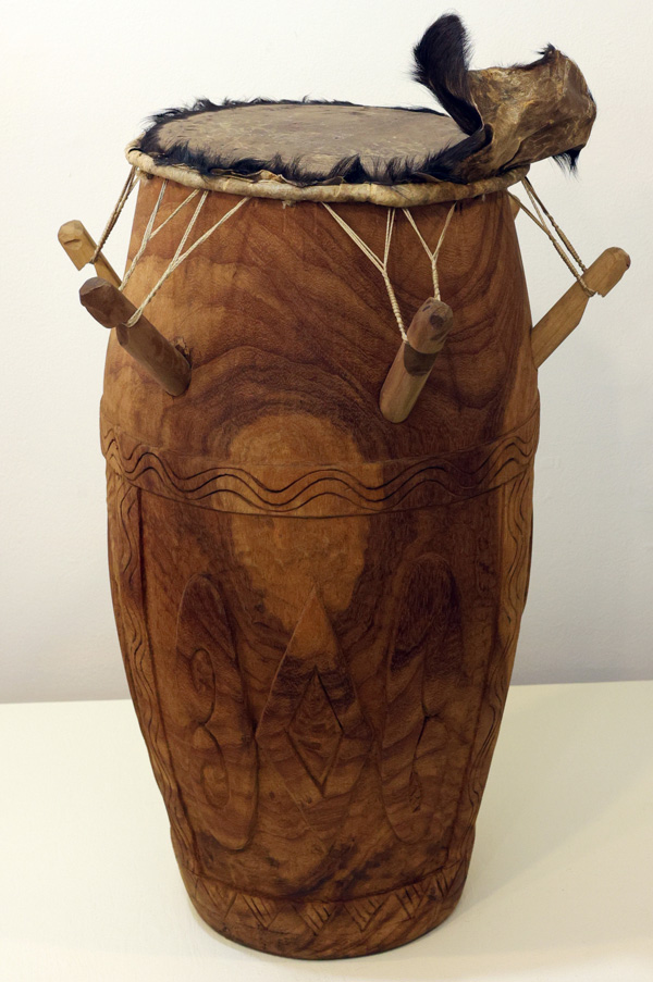 Image of a tall brown wooden drum with details carved into the middle and bottom of the drum. The drum has four wooden sticks extending out of the upper half with thin white sting connecting them to the top of the drum. The top of the drum has a white rim followed by a fuzzy black rim on top of it and a gray head. A black and brown piece extends off of the top of the rim on the right side of the image.