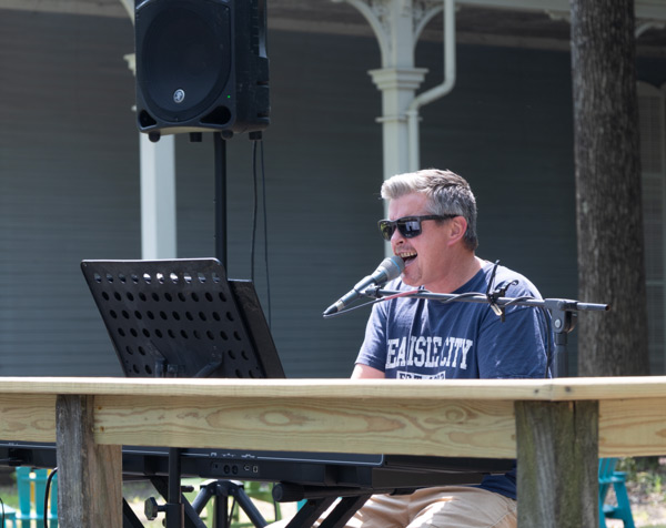 Image of Dale Bierman sitting outside on a wooden stage next to the WheatonArts Museum of American Glass. Dale is singing into a microphone and is using both hands to play the large keyboard that is in front of him. There is a tall speaker in the background on the left side of the image.