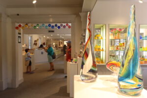 Image of the inside of the WheatonArts Paperweight Shop. On the right side of the image are two tall clear glass statues that are wide and round at the bottom before turning thin at the top. The statue on the left has a rainbow swirl in the center and the statue on the right has a blue and yellow swirl. Both statues sit on the top of a white podium and the back of a rainbow glass heart is visible behind the statues on the same podium. In the background, there are three cases inside of the white wall filled with glass pumpkins, glass vases, and glass cups resting on the shelves. Each case has three shelves. To the left of these cases is a black hanging rack filled with glass ornaments and two smaller podiums with glass paperweights on top of them. To the left of these is the entryway to the Gallery of Fine Craft. The entryway has colorful glass ornaments hanging from the top in rainbow order. Two adults and a child can be seen past the entryway. One of the adults is takin a photo of the child posing next to a hand-carved white wooden egret that is resting on top of a podium.