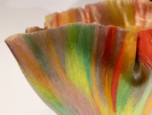 Close up image of a piece by Toots Zynsky. The image shows the left side of a colorful bowl-shaped glass pieces that folds and ruffles at the top. The piece is beige with bright light green, green, yellow, orange, and red thick vertical streaks throughout