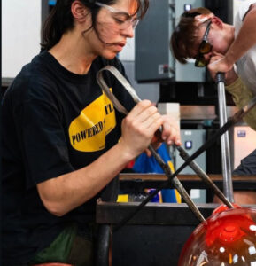 Side profile image of Anna Antonia Yiaxi Savvidou working at a glassblowing bench. Anna is using both hands to shape hot glass with a large pair of metal tweezers. A small corner of the hot glass is pictured at the bottom right of the image.