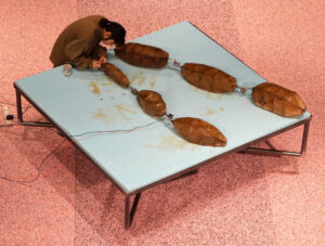 Image of Anna Antonia Yiaxi Savvidou working at a large white square table. There is a large art piece resting on top of it. The piece is comprised of six large amber colored cacoon-like pieces connected together in a long strand with metal hooks. The artist is working near one of the hooks at the edge of the table. One of the hooks also has long thin wires connected to it.