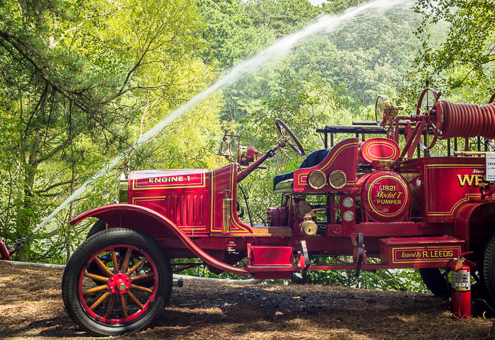 Image of a red antique fire truck outside in front of the WheatonArts lake at Fire Muster. There is a hose on the left side of the truck that is shooting a stream of water in an arc into the lake.