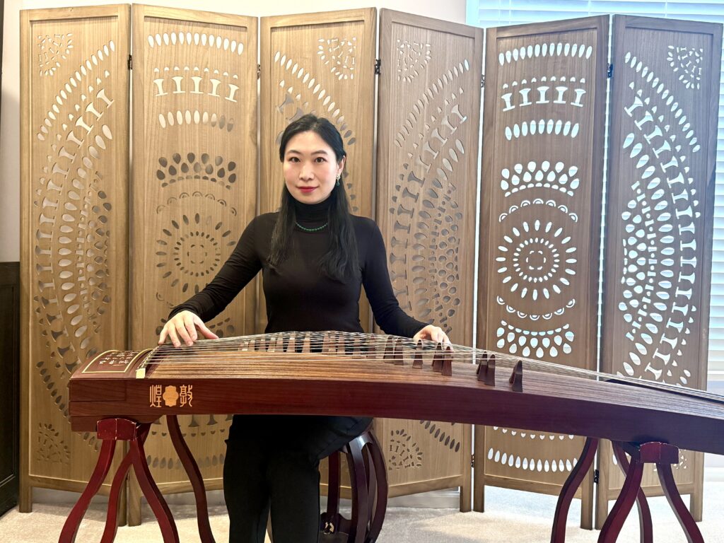 Image of April Yang sitting behind a guzheng with both hands on the strings. There is a 6-panel wooden room divider behind April with hand carved cutouts that create two circular designs. One is on the left 3 panels and one is on the right 3 panels.