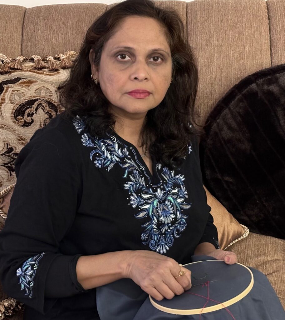 Image of Shikha Dwivedi sitting with an embroidery hoop with black fabric inside of it. Shikha is using red thread and a needle to create the beginning of a larger design on the fabric inside of the hoop.