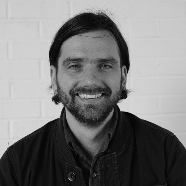 Grayscale headshot image of Charles Manion. Charles has dark hair that falls below the ears and a short beard.