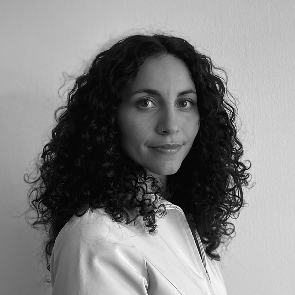 Grayscale headshot image of Felicity Machado. Felicity has dark, curly hair that falls below the shoulders. Felicity is turned to the side with her head turned towards the camera.